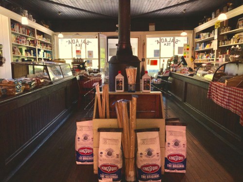 A cozy store interior with wooden shelves, various products, and a central stove, featuring bags of charcoal in the foreground.