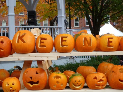 A display of carved pumpkins on a wooden stand, featuring the name "KEENE" and various playful faces.