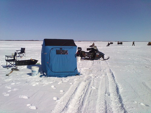 A blue ice fishing shelter on a snowy landscape, with snowmobiles and chairs in the background under a clear sky.