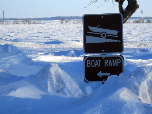 A snow-covered landscape with a sign indicating a boat ramp, pointing left amidst the winter scenery.
