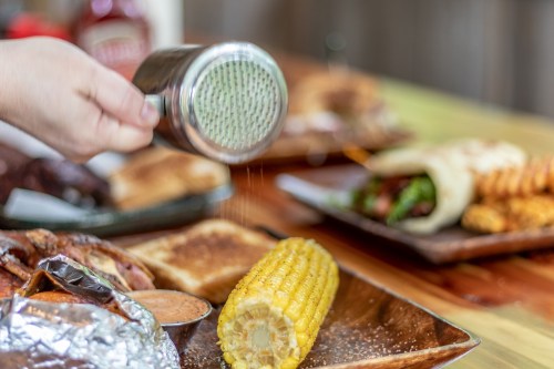 A hand shakes a spice shaker over a cob of corn, with various dishes in the background on a wooden table.