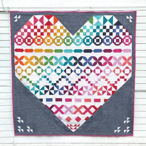 A colorful quilt in the shape of a heart, featuring various geometric patterns and a gray background.
