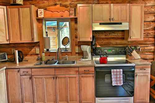 Cozy log cabin kitchen with wooden cabinets, a stove, sink, and a red kettle on the counter. Natural light from the window.