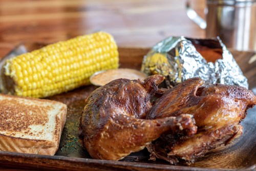 A wooden tray with grilled chicken, corn on the cob, a baked potato in foil, toast, and dipping sauce.
