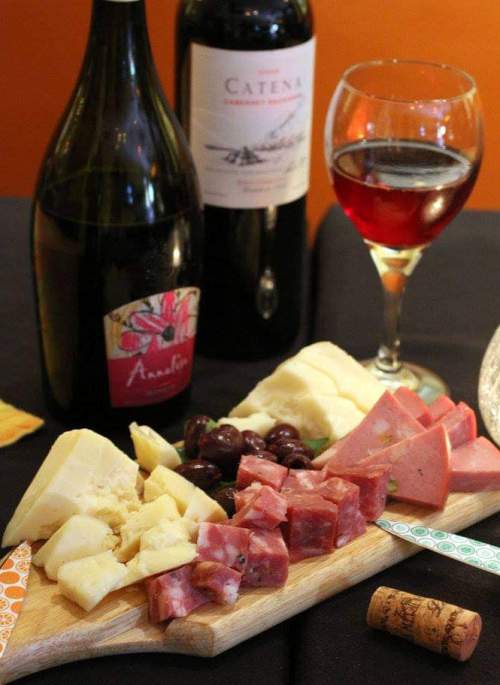 A wooden board with assorted cheeses, meats, and olives, accompanied by two bottles of wine and a glass of red wine.