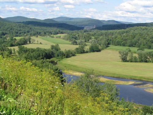 A scenic landscape featuring rolling hills, a river, and lush greenery under a partly cloudy sky.