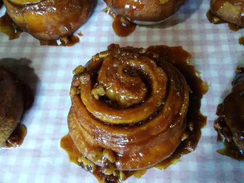 Close-up of a sticky, caramel-coated cinnamon roll topped with chopped nuts on a checkered paper background.