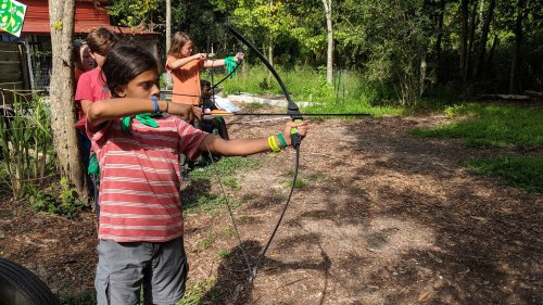 A child aims a bow and arrow in a wooded area, with others practicing archery in the background.