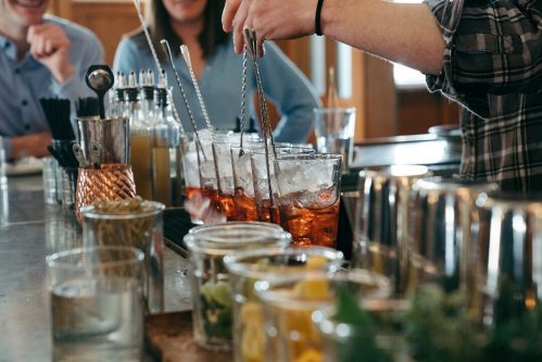 A bartender prepares cocktails with ice and garnishes at a lively bar, with patrons enjoying drinks in the background.