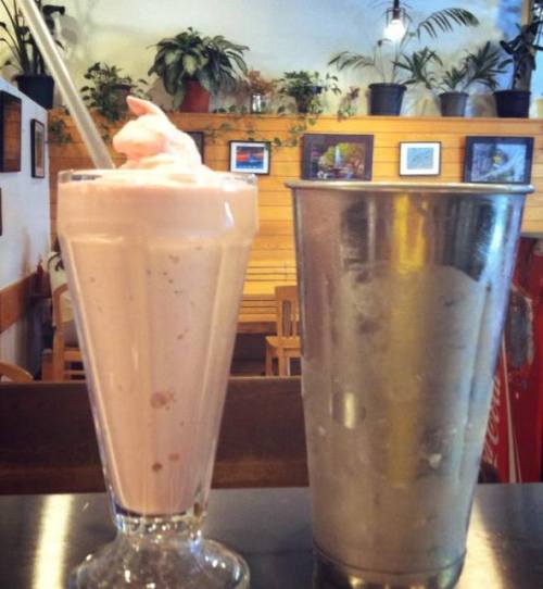 A tall glass of pink milkshake with whipped cream next to a metal cup, both set in a cozy café with plants in the background.