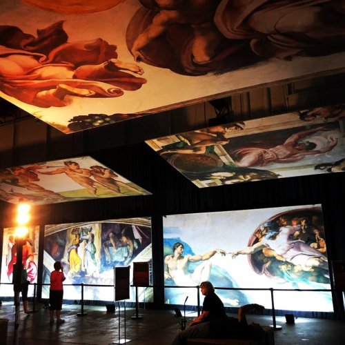 Visitors admire large-scale reproductions of Michelangelo's ceiling paintings in a dimly lit exhibition space.