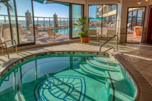 A hot tub with a decorative design, overlooking a pool area and ocean view through large windows.