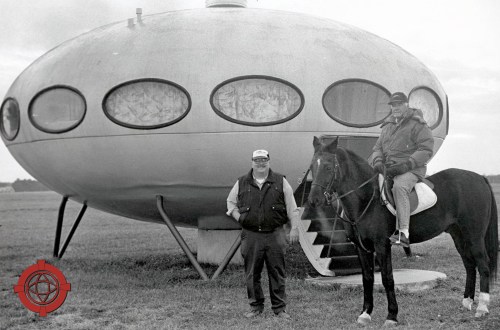 Two men stand beside a futuristic, oval-shaped building, one on a horse, in a grassy field.