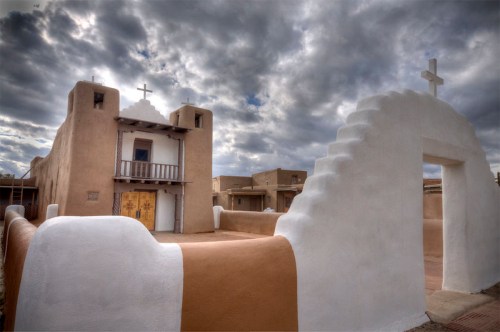 A historic adobe church with crosses, featuring a white and brown exterior under a cloudy sky.