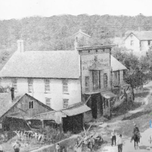 Historic black-and-white image of a building with a sign, surrounded by trees and a dirt road with people and horses.