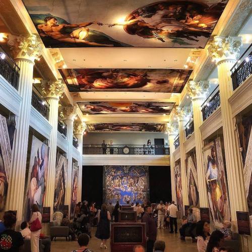 A grand museum interior with high ceilings, ornate columns, and large murals, filled with visitors admiring the art.