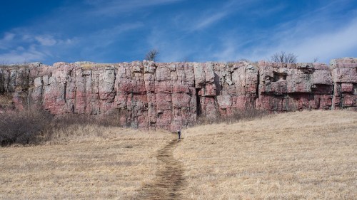 A person walks along a path toward a large, colorful rock formation under a blue sky with scattered clouds.