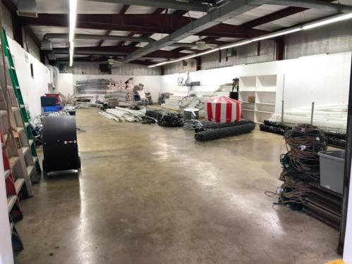 A spacious, empty warehouse with scattered equipment, shelves, and a ladder against the wall.