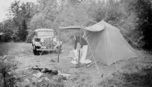A vintage black-and-white photo of a tent and an old car in a grassy area surrounded by trees.