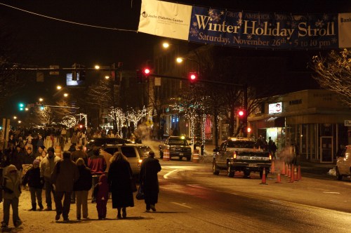 A busy street at night with holiday decorations, people walking, and a banner for a Winter Holiday Stroll event.