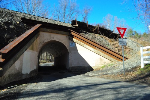 A road passes under a stone archway with a yield sign, surrounded by trees and gravel.