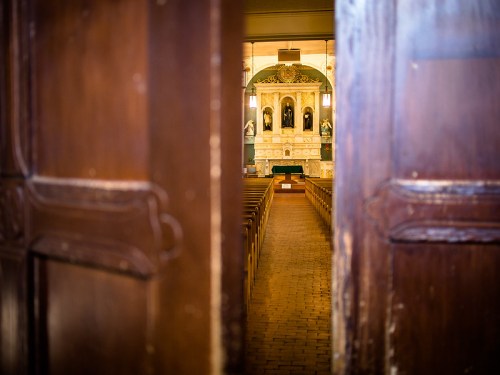 View through open wooden doors into a church interior, featuring rows of pews and an ornate altar at the front.