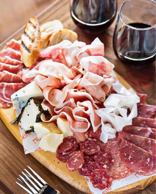 A wooden platter filled with assorted meats, cheeses, and bread, accompanied by two glasses of red wine.