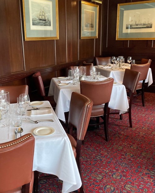 Elegant restaurant interior with wooden walls, framed ship paintings, and neatly set tables with white tablecloths.