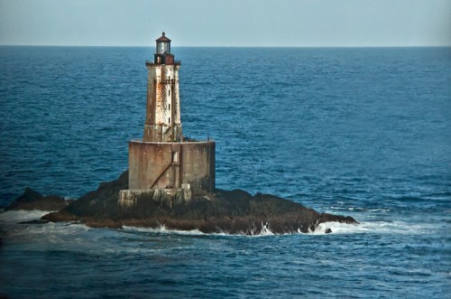 A weathered lighthouse stands on a rocky outcrop, surrounded by calm blue ocean waters.