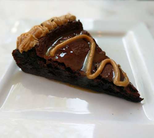 A slice of rich chocolate cake drizzled with caramel and topped with a creamy peanut butter swirl on a white plate.