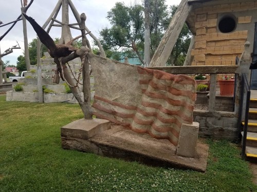 A weathered sculpture of a flag draped over a stone bench, set in a grassy area with trees and a building in the background.