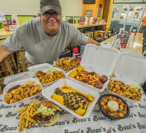 A smiling man sits at a table surrounded by various seafood dishes, burgers, fries, and takeout containers.