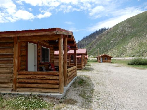 Log cabins with red roofs sit along a gravel path, surrounded by green hills and a blue sky with scattered clouds.