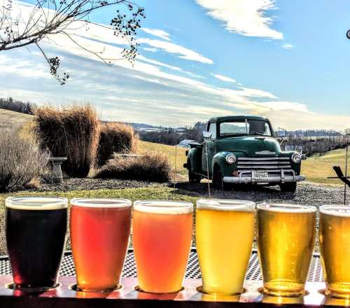 A row of colorful craft beers in front of a vintage green truck and a scenic landscape under a blue sky.