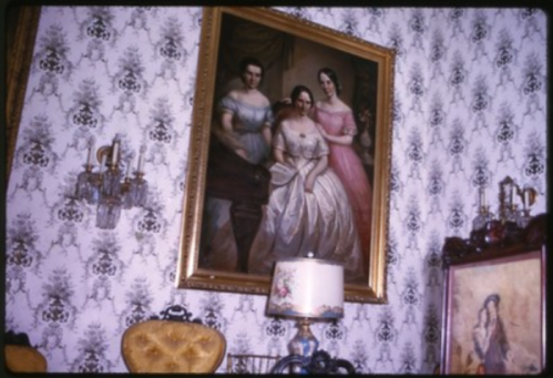 A vintage room featuring a large painting of three women, ornate wallpaper, and antique furniture.