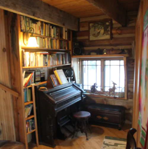 Cozy wooden interior featuring a bookshelf, an old piano, and a window with a view, creating a warm atmosphere.