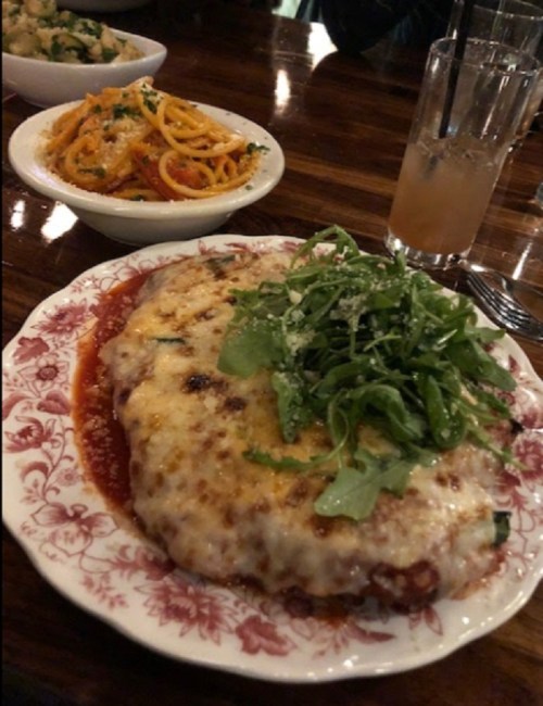 A plate of cheesy chicken parmesan topped with arugula, served with a side of spaghetti and a drink in the background.