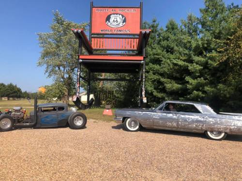 Two classic cars parked under a Route 66 sign, surrounded by trees and gravel. One car is vintage with flames, the other is wrapped in foil.