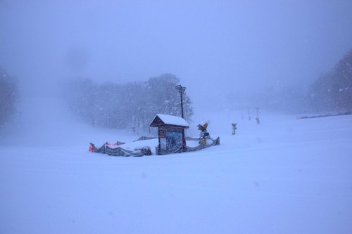 A snowy landscape with a small building and ski lift in the distance, surrounded by heavy snowfall.