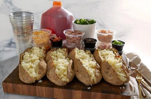 Three baked potatoes on a wooden board, surrounded by various toppings in containers, including cheese, meats, and greens.