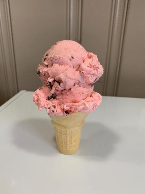 A cone of pink ice cream with chocolate chunks, topped with a generous scoop, set against a neutral background.