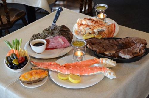 A table spread with a variety of meats, including prime rib, crab legs, and sides of vegetables and baked potato.