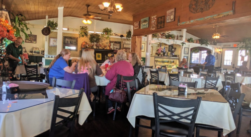 A cozy restaurant interior with tables set for dining, featuring patrons chatting and a server attending to guests.
