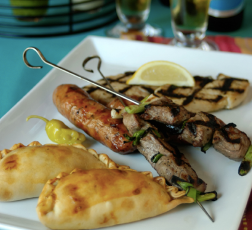 A plate of grilled meats, including skewers and sausages, alongside golden empanadas and a lemon wedge.