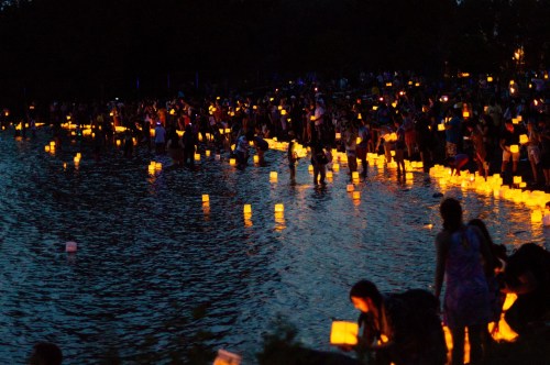 A crowd gathers by a lake at dusk, releasing glowing lanterns into the water, creating a serene, illuminated scene.