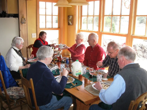 A group of seven people enjoying a meal together at a sunny dining table with large windows.