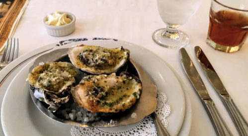 A plate of baked oysters topped with herbs, served on a bed of salt, with a glass of water and a drink in the background.