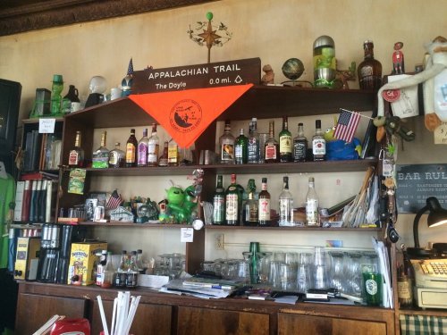 A wooden bar shelf filled with various liquor bottles, glasses, and decorations, including an orange Appalachian Trail flag.