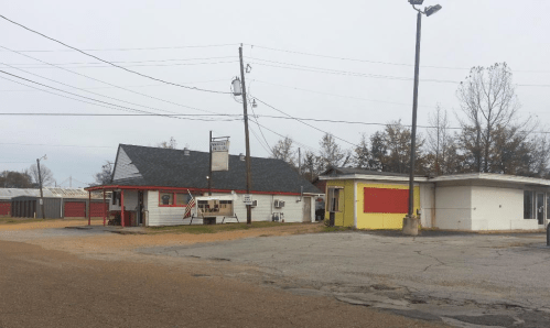 A small, single-story building with a red and white exterior, next to a vacant yellow building on a gravel road.