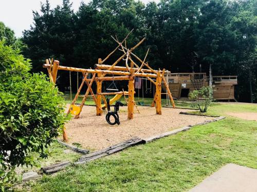 A wooden playground with swings and a climbing structure, surrounded by grass and trees. A playhouse is in the background.